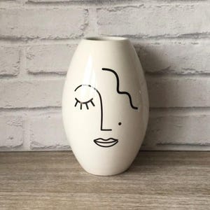 May include: Cream ceramic vase with a minimalist face design. The face is outlined in black, featuring a closed eye, a nose, a mouth, and a wavy hair design. The vase has a smooth, glossy finish and a rounded, oval shape.