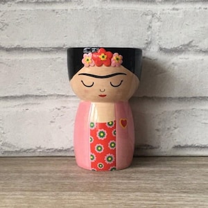 以下が含まれることがあります： 女性の顔と胴体を模した手描きのセラミックプランター。 プランターは黒い上部、目を閉じた顔、花柄のピンク色のドレスが特徴です。 ドレスにはハートが描かれています。