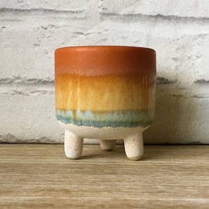 May include: A small, ceramic planter with a gradient of colours, including orange, yellow, and blue. The planter has three short, rounded legs and a textured surface. The design is reminiscent of a sunset. Ideal for small plants.
