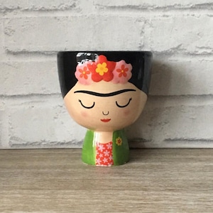 以下が含まれることがあります： 顔の形をしたセラミックプランター。黒い上部、閉じた目、花の冠が特徴です。プランターは、赤と白の模様のドレスの上に緑色のジャケットを着ています。顔は明るい肌の色で塗装されています。