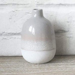 Puede incluir: Un pequeño jarrón decorativo con cuerpo redondeado y cuello estrecho. El jarrón está pintado en degradado de colores, pasando del blanco en la base al gris claro en la parte superior, con una banda rosa pálido en el medio. El jarrón es de cerámica.