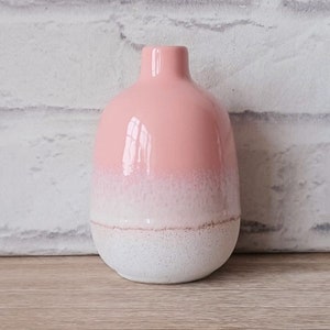 Op de afbeelding: Een kleine, roze en witte keramische vaas met een getextureerde, witte basis. De vaas heeft een ronde, bolle vorm en een smalle hals.