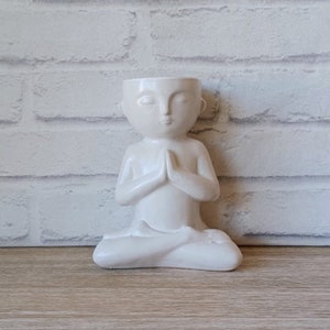 Puede incluir: Maceta de cerámica blanca con forma de persona sentada en una postura de yoga meditativa con las manos juntas frente al pecho. La maceta tiene una cabeza hueca que se puede utilizar para sostener una planta pequeña.