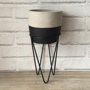 May include: A white and black planter with a black metal stand. The stand has a triangular shape with three legs.