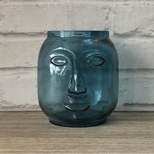 Pode incluir: Um vaso de vidro azul-petróleo com um design de rosto. O vaso tem uma forma arredondada com uma abertura larga e apresenta um rosto esculpido com olhos, nariz e boca. O vaso está sobre uma superfície de madeira.