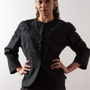 Può includere: Blazer nero con ricami floreali decorativi sulle spalle e sulla parte superiore del petto. Il blazer ha un colletto rotondo, tre bottoni e maniche a tre quarti. La modella indossa pantaloni neri.