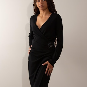 May include: A black wrap dress with long sleeves and a V-neck. The dress features a decorative embellishment at the waist. The model is wearing rings. The dress is knee-length.