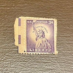 May include: A vintage 3-cent U.S. postage stamp featuring the Statue of Liberty, with the text "GOD WE TRUST" and "LIBERTY". The stamp is purple and tan, with perforated edges.