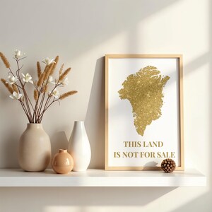 Peut inclure: Un cadre avec une carte en paillettes dorées d'une masse terrestre, avec le texte "THIS LAND IS NOT FOR SALE". Le cadre est posé sur une étagère blanche avec des vases et des fleurs décoratives.