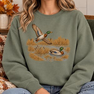 Peut inclure: Un sweat-shirt à col rond vert olive orné d'une broderie représentant deux canards colverts dans un marais. Un canard est représenté en plein vol, ailes déployées, tandis que l'autre nage dans l'eau parmi de grands roseaux dorés.