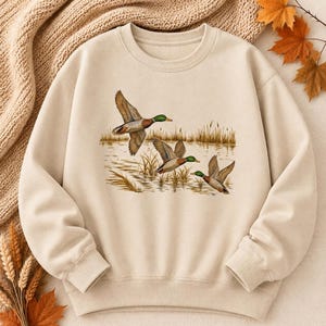 Peut inclure: Un sweat-shirt à col rond beige avec une impression graphique centrale représentant trois canards colverts en vol au-dessus d'un étang marécageux avec de hautes herbes. Le motif montre les canards à tête verte sur un fond en tissu beige.