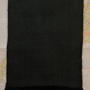 Puede incluir: Bufanda negra con un patr&oacute;n de espiga y flecos en la parte inferior. El patr&oacute;n de la bufanda es una V repetida. La bufanda est&aacute; hecha de material tejido y es de color negro liso.