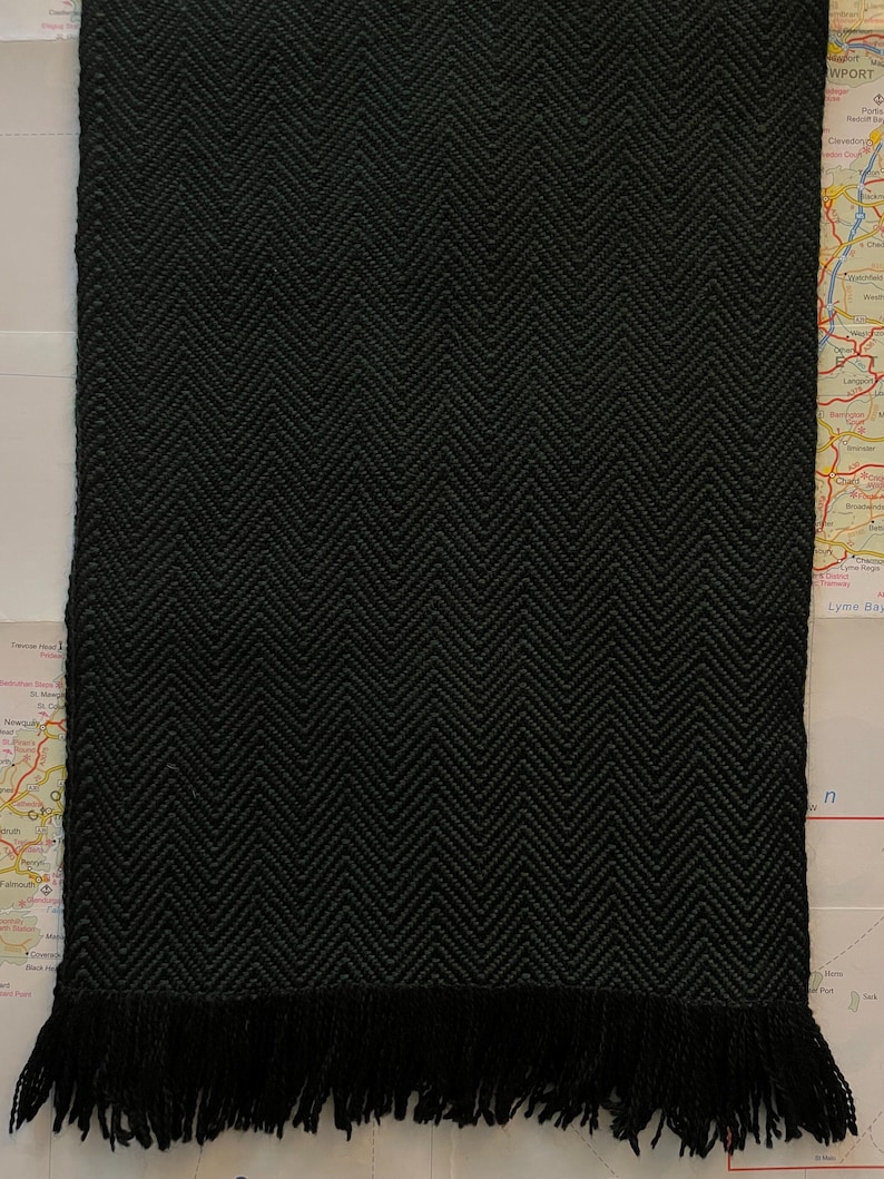 Puede incluir: Bufanda negra con un patr&oacute;n de espiga y flecos en la parte inferior. El patr&oacute;n de la bufanda es una V repetida. La bufanda est&aacute; hecha de material tejido y es de color negro liso.