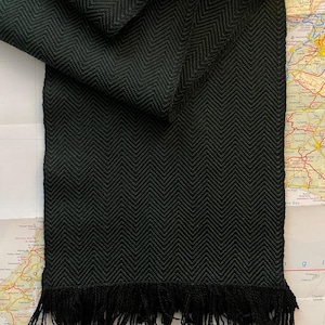 Puede incluir: Bufanda verde oscuro con un patr&oacute;n de espiga tejido y flecos. La bufanda est&aacute; doblada sobre un fondo de mapa, mostrando la textura y el dise&ntilde;o. Los flecos est&aacute;n en la parte inferior de la bufanda.