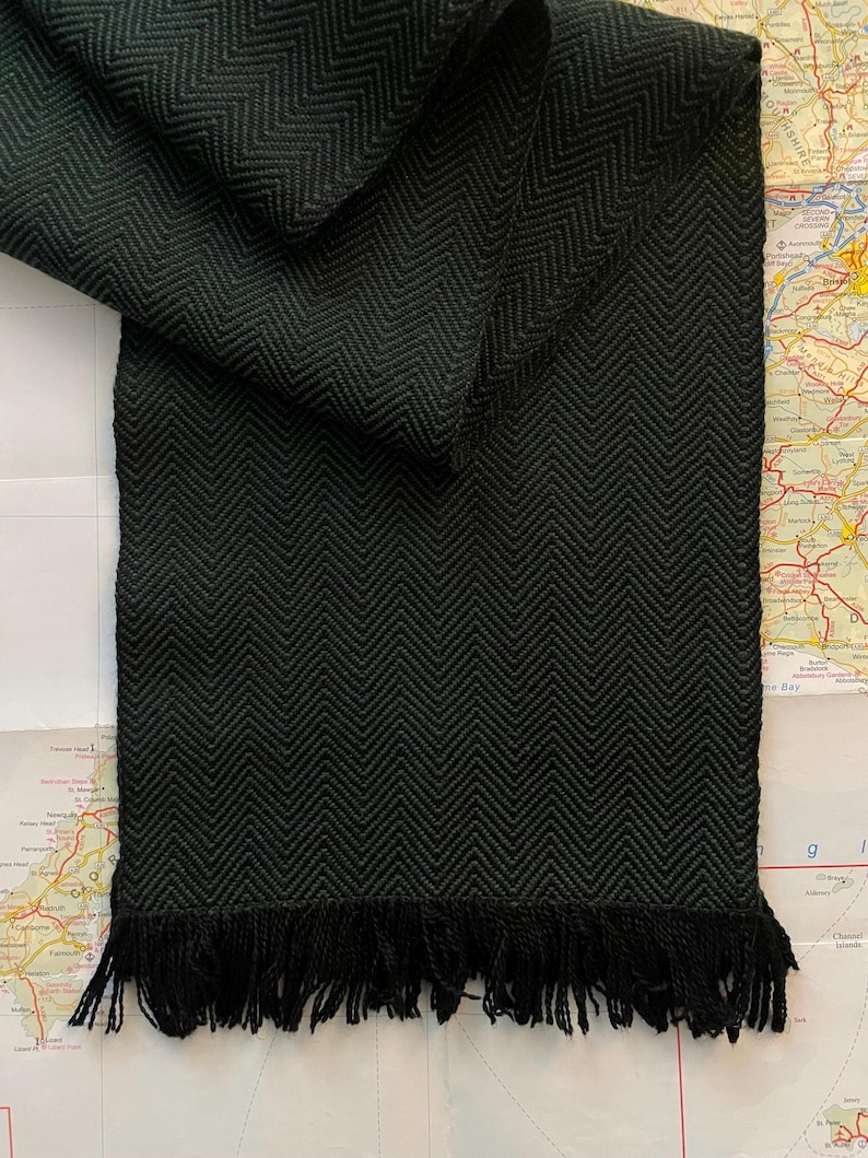 Puede incluir: Bufanda verde oscuro con un patr&oacute;n de espiga tejido y flecos. La bufanda est&aacute; doblada sobre un fondo de mapa, mostrando la textura y el dise&ntilde;o. Los flecos est&aacute;n en la parte inferior de la bufanda.