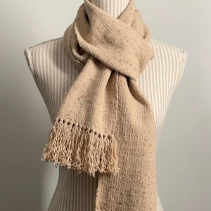 May include: A beige scarf with fringe detail, displayed on a mannequin. The scarf is made of a woven fabric with a textured appearance. The fringe hangs at the ends of the scarf. The scarf is draped around the mannequin's neck.