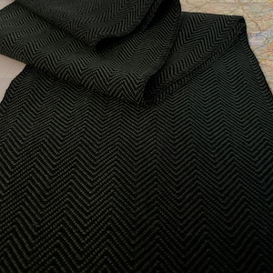 Puede incluir: Una bufanda verde oscuro con un patr&oacute;n de espiga negro. La bufanda est&aacute; doblada sobre una superficie de color claro, mostrando el intrincado tejido y la textura. La bufanda parece estar hecha de un material suave y c&aacute;lido.