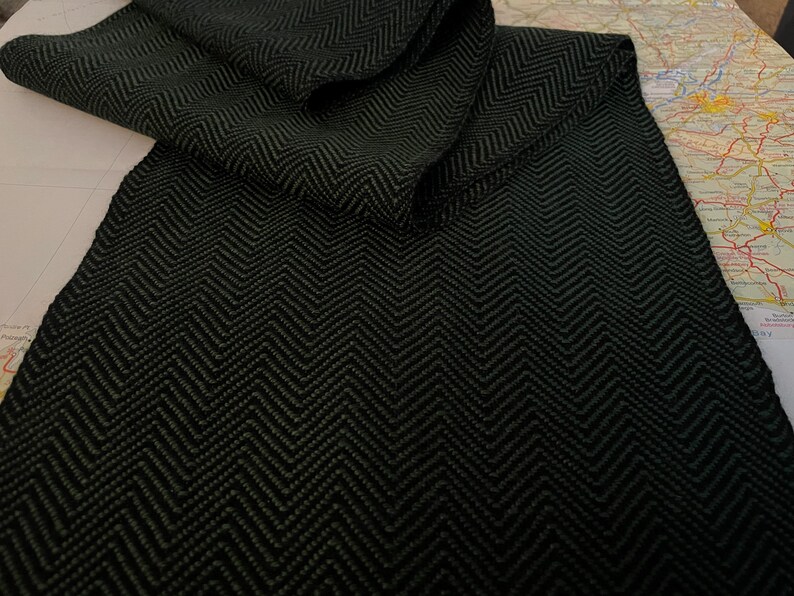 Puede incluir: Una bufanda verde oscuro con un patr&oacute;n de espiga negro. La bufanda est&aacute; doblada sobre una superficie de color claro, mostrando el intrincado tejido y la textura. La bufanda parece estar hecha de un material suave y c&aacute;lido.