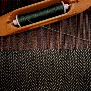 Puede incluir: Primer plano de una lanzadera de madera que sostiene hilo verde oscuro, colocada sobre una tela tejida con un patr&oacute;n de espiga en blanco y negro. La tela est&aacute; parcialmente terminada, mostrando el proceso de tejido.