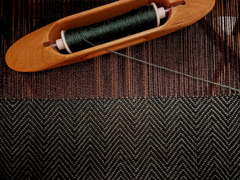 Puede incluir: Primer plano de una lanzadera de madera que sostiene hilo verde oscuro, colocada sobre una tela tejida con un patr&oacute;n de espiga en blanco y negro. La tela est&aacute; parcialmente terminada, mostrando el proceso de tejido.