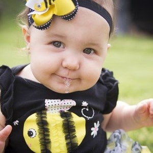 Puede incluir: Una ni&ntilde;a peque&ntilde;a con un atuendo negro y amarillo con tem&aacute;tica de abejas y un lazo amarillo en el pelo. Est&aacute; sentada en una superficie verde cubierta de hierba y tiene un poco de baba en la barbilla.