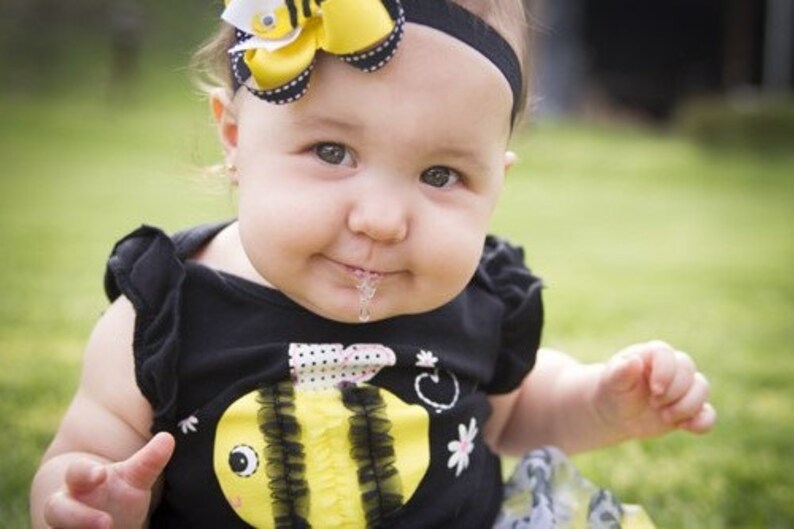 Puede incluir: Una ni&ntilde;a peque&ntilde;a con un atuendo negro y amarillo con tem&aacute;tica de abejas y un lazo amarillo en el pelo. Est&aacute; sentada en una superficie verde cubierta de hierba y tiene un poco de baba en la barbilla.
