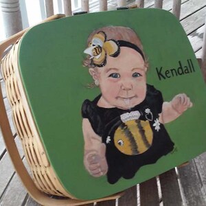 Puede incluir: Una cesta de picnic de mimbre verde con una imagen pintada de una ni&ntilde;a peque&ntilde;a con una diadema con tem&aacute;tica de abeja y un vestido negro con un dise&ntilde;o de abeja. La cesta tiene el nombre "Kendall" pintado en el lateral.