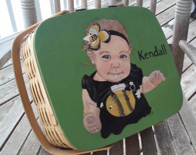 Puede incluir: Una cesta de picnic de mimbre verde con una imagen pintada de una ni&ntilde;a peque&ntilde;a con una diadema con tem&aacute;tica de abeja y un vestido negro con un dise&ntilde;o de abeja. La cesta tiene el nombre "Kendall" pintado en el lateral.