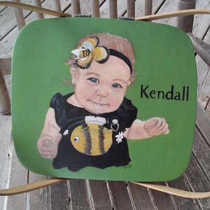 Puede incluir: Una cesta de picnic de madera verde con un retrato pintado de una ni&ntilde;a peque&ntilde;a que lleva un vestido negro y una diadema con tem&aacute;tica de abeja. La ni&ntilde;a sostiene un juguete con forma de abeja. La cesta est&aacute; etiquetada con "Kendall".