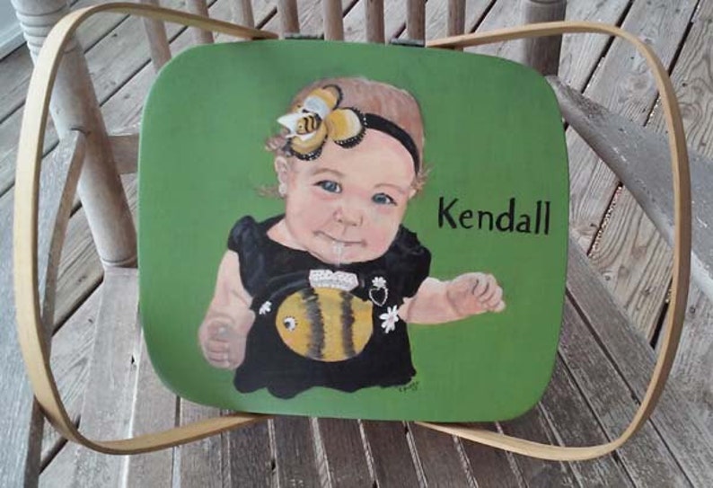 Puede incluir: Una cesta de picnic de madera verde con un retrato pintado de una ni&ntilde;a peque&ntilde;a que lleva un vestido negro y una diadema con tem&aacute;tica de abeja. La ni&ntilde;a sostiene un juguete con forma de abeja. La cesta est&aacute; etiquetada con "Kendall".