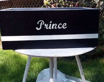 May include: Black wooden planter box with a white stripe and the word "Prince" painted in white cursive lettering.