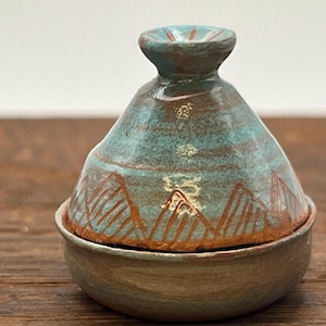 Puede incluir: Pequeño tajín artesanal de cerámica con esmalte turquesa y marrón. La tapa tiene forma cónica con un pequeño asa, y la base es redonda. La olla está decorada con un diseño de montaña, ideal para cocinar y servir.