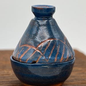 Puede incluir: Un tarro de cerámica azul oscuro hecho a mano con tapa. El tarro presenta un diseño único de líneas marrones en la superficie. El tarro está sobre una superficie de madera. Un artículo decorativo.