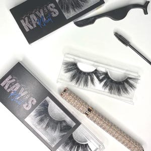 May include: An overhead shot of beauty products, including false eyelashes, eyelash applicators, and a mascara wand. The image features a box with the text "Kay's Kloset" in silver glitter and blue script. The eyelashes are in a clear plastic case.