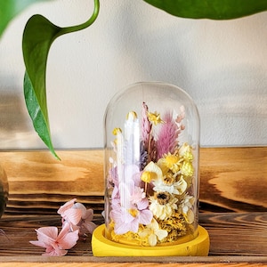 Peut inclure: Un dôme en verre avec une base jaune repose sur une étagère en bois. À l'intérieur du dôme, des fleurs séchées dans des tons de rose, jaune et blanc.