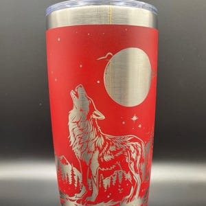 May include: A red stainless steel tumbler featuring a laser-etched design of a wolf howling at the moon. The wolf is silhouetted against a red background with a silver moon and stars.
