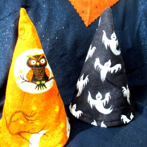 May include: Two Halloween-themed cone hats. One is orange with an owl and moon design. The other is black with white ghost patterns. Both are made of fabric and are likely for decoration or costume.