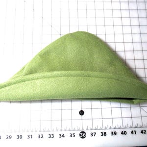 May include: A light green, triangular-shaped hat with a rolled brim. The hat is made of a soft, felt-like material. The hat is resting on a white surface with a grid pattern and measurement markings.