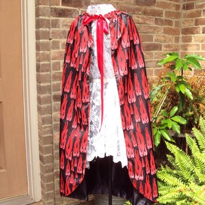 May include: A black cape with a red flame pattern, a red ribbon, and a white lace garment. The cape is displayed outdoors against a brick wall and greenery. The cape is a costume or theatrical garment.