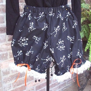 May include: Black shorts with a white skeleton print. The shorts have a ruffled white lace trim and orange ribbon ties. The elastic waistband is visible. The shorts are likely for Halloween or a costume.