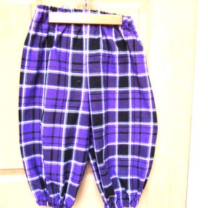 May include: Purple, black, and white plaid pants with a gathered waist and ankles. The pants are hanging on a wooden hanger against a light-colored background. The fabric has a soft, slightly textured appearance.