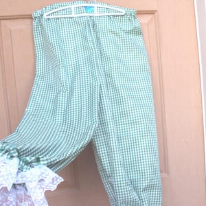 May include: Green and white gingham patterned pants with elastic cuffs and white lace trim. The pants are hanging on a white hanger against a light brown door.