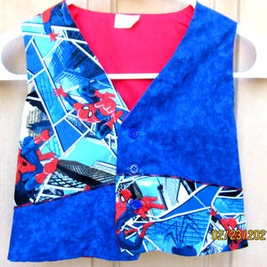 May include: A blue and red Spider-Man themed vest with a V-neck design. The vest features a vibrant red lining and is adorned with images of Spider-Man in action against a cityscape backdrop. It has blue buttons and is hanging on a white hanger.