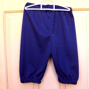 May include: Navy blue capri trousers with an elasticated waistband and gathered cuffs. The trousers are hanging on a white hanger against a light-coloured wooden door.