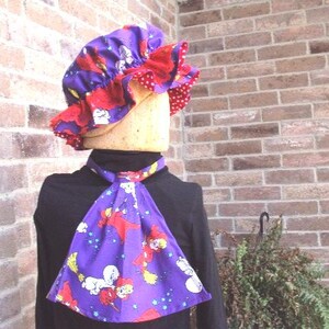 May include: A purple bonnet and scarf set featuring a cartoon character print. The bonnet has a red ruffled trim and a red polka dot lining. The scarf is triangular and has the same cartoon character print as the bonnet.