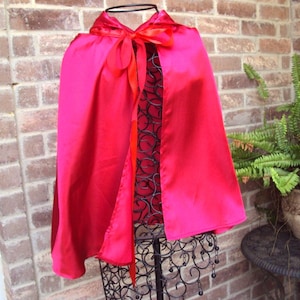 May include: A bright pink satin cape with a hood and a red bow at the neck. The cape is displayed on a black metal stand against a brick wall, with a potted fern in the background.