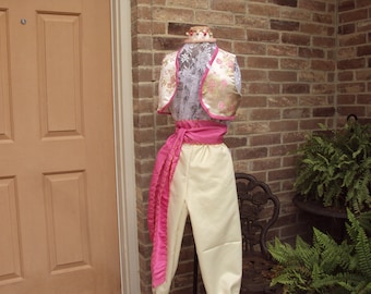 May include: A white and gold brocade vest with pink trim and a pink sash. The vest is worn over a white lace top and the sash is tied at the waist. The outfit is displayed on a mannequin against a brick wall.