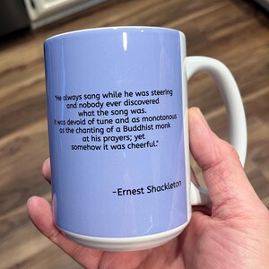 May include: A white ceramic mug with a light blue background and black text. The text reads: "He always sang while he was steering and nobody ever discovered what the song was. It was devoid of tune and as monotonous as the chanting of a Buddhist monk at his prayers; yet somehow it was cheerful." -Ernest Shackleton