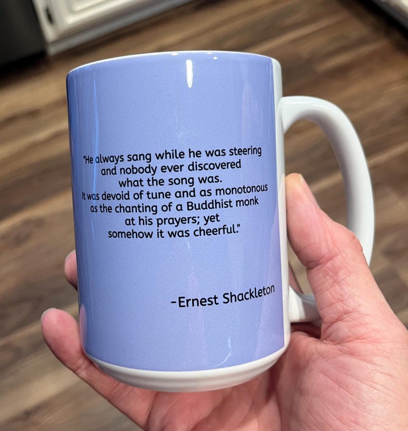 May include: A white ceramic mug with a light blue background and black text. The text reads: "He always sang while he was steering and nobody ever discovered what the song was. It was devoid of tune and as monotonous as the chanting of a Buddhist monk at his prayers; yet somehow it was cheerful." -Ernest Shackleton