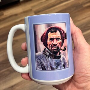 May include: A white ceramic mug with a blue background and a framed image of a man with a beard wearing a red hat and a pipe. The man is looking directly at the viewer.